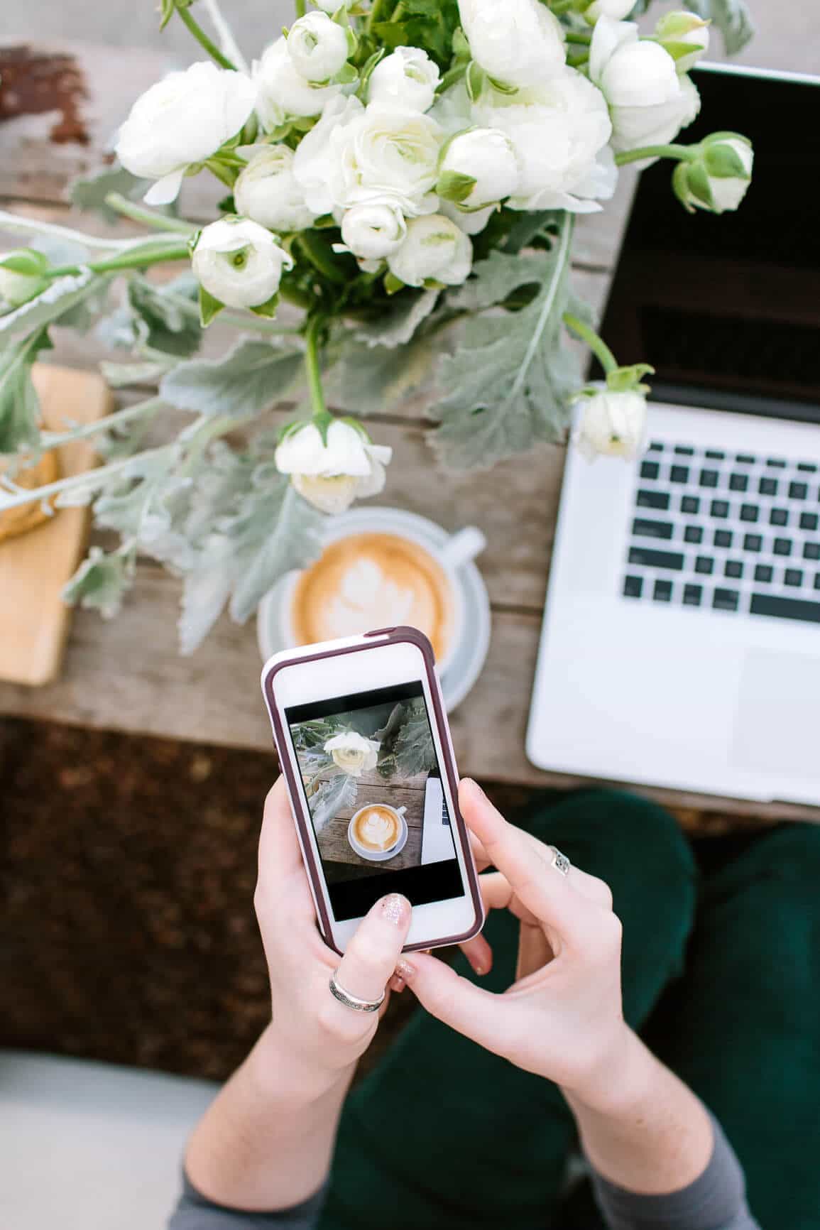 Woman taking iphone photo of her coffee on beautiful table with flowers and laptop.